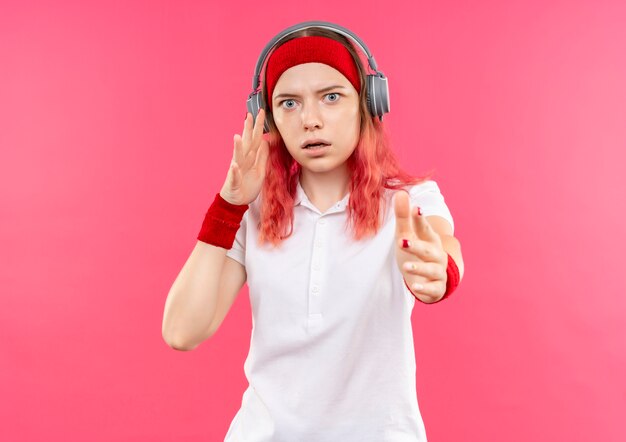 Jeune femme sportive en bandeau avec un casque surpris debout sur un mur rose