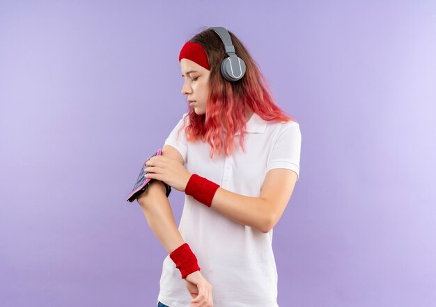 Jeune femme sportive en bandeau avec des écouteurs touchant son brassard de smartphone debout sur un mur violet