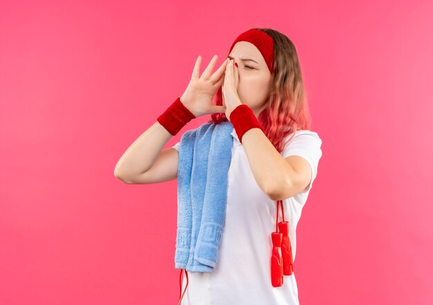 Jeune femme sportive en bandeau avec une serviette sur l'épaule appelant quelqu'un avec des paumes près de la bouche debout sur un mur rose