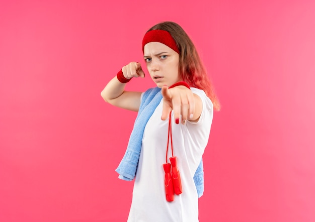 Jeune femme sportive en bandeau avec une serviette sur l'épaule pointant avec le doigt à la caméra avec un visage sérieux debout sur un mur rose