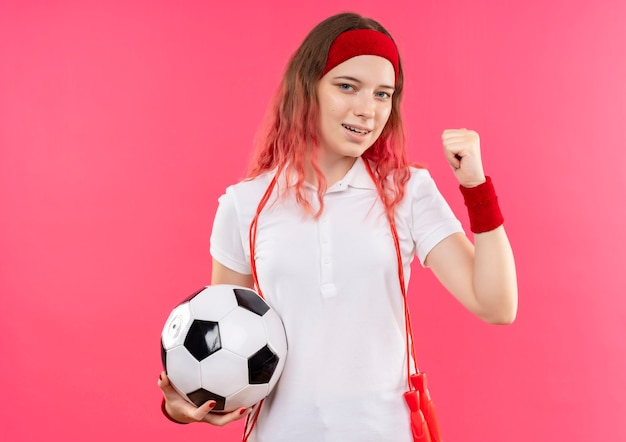 Jeune femme sportive en bandeau tenant un ballon de football serrant le poing heureux et sorti debout sur un mur rose