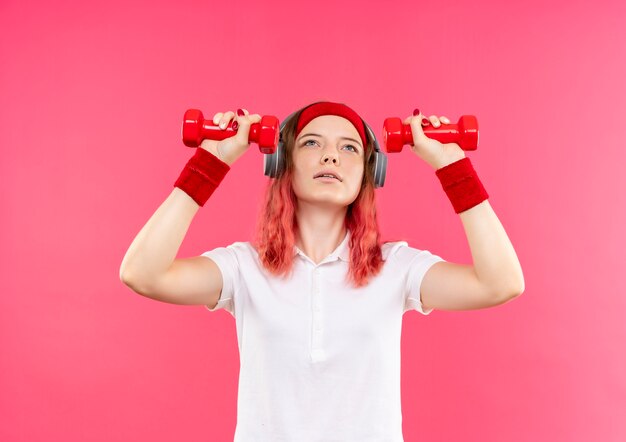 Jeune femme sportive en bandeau tenant deux haltères faisant des exercices debout sur un mur rose