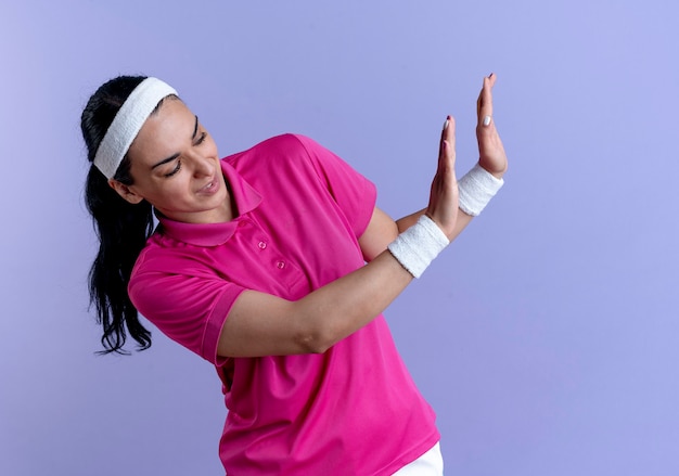 Jeune femme sportive caucasienne agacée portant un bandeau et des bracelets fait semblant de pousser quelque chose à la recherche de côté sur violet avec copie espace