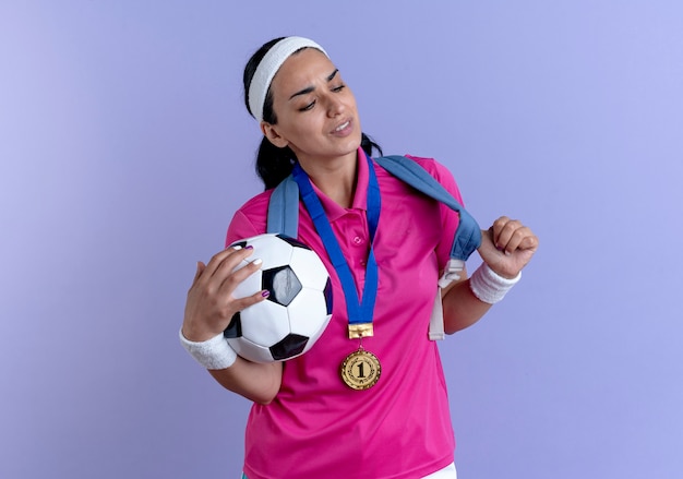 Jeune femme sportive caucasienne agacée portant bandeau de sac à dos et bracelets avec médaille d'or autour de son cou tient balle isolée sur espace violet avec espace copie