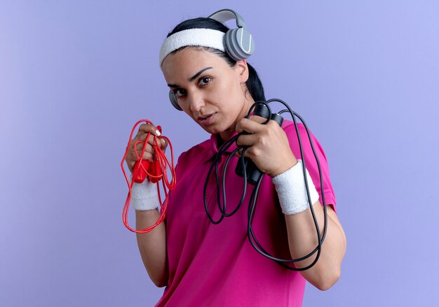 Jeune femme sportive caucasienne confiante portant bandeau et bracelets sur les écouteurs détient des cordes à sauter isolé sur fond violet avec copie espace