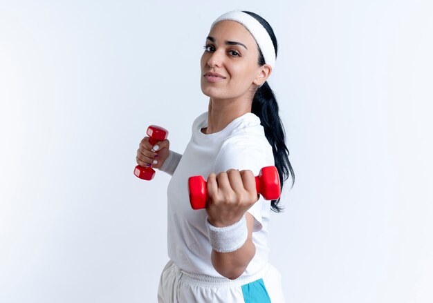 Jeune femme sportive caucasienne confiante portant bandeau et bracelets se tient sur le côté tenant haltère isolé sur un espace blanc avec copie espace