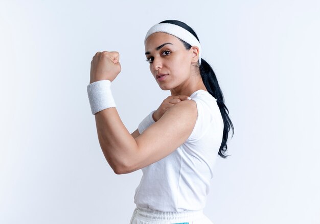 Jeune femme sportive caucasienne confiante portant bandeau et bracelets se tient sur le côté et tend les biceps isolés sur un espace blanc avec copie espace