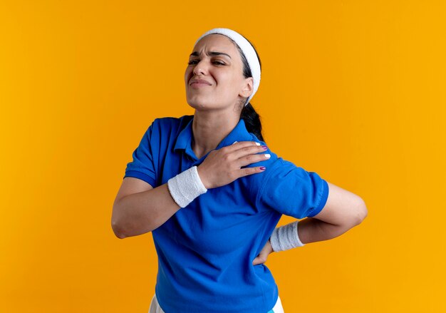 Jeune femme sportive caucasienne endolori portant bandeau et bracelets tient l'épaule et le dos isolé sur fond orange avec copie espace