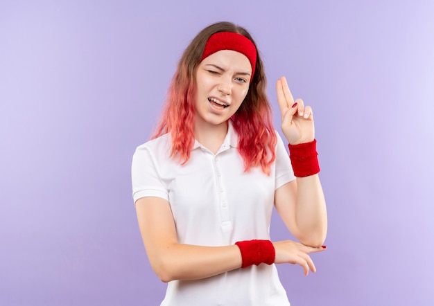 Jeune femme sportive un clin de œil et souriant montrant deux doigts debout sur un mur violet