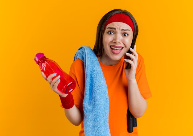 Jeune femme sportive en colère portant bandeau et bracelets avec corde à sauter et serviette sur les épaules tenant une bouteille d'eau et parler au téléphone à tout droit