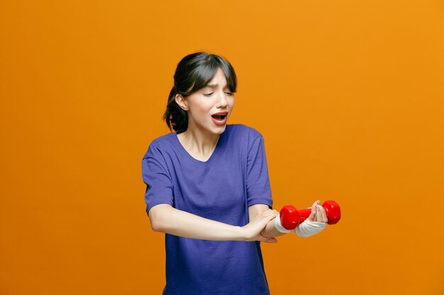 Jeune femme sportive endolorie portant un t-shirt saisissant son bras tenant une haltère en le regardant avec un poignet enveloppé de bandage isolé sur fond orange