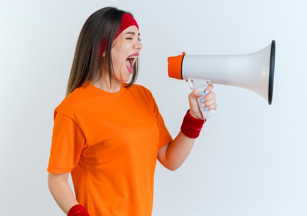 Jeune femme sportive portant bandeau et bracelets debout en vue de profil en criant dans le haut-parleur avec les yeux fermés