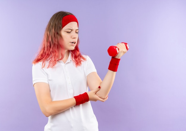 Jeune femme sportive tenant un haltère à faire des exercices en regardant la douleur au coude debout sur le mur violet