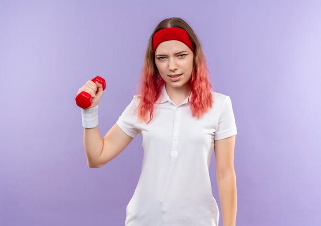Jeune femme sportive tenant des haltères à la main avec un visage sérieux debout sur un mur violet