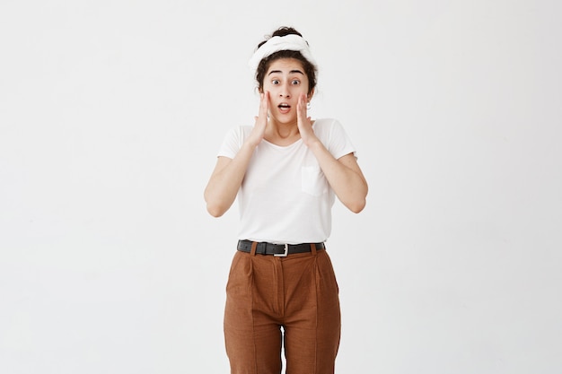 Jeune femme stupéfaite choquée en t-shirt blanc et pantalon marron regarde avec les yeux sombres et la bouche ouverte, garde les mains sur les joues, ne croit pas en son échec, a perplexe regard frustré