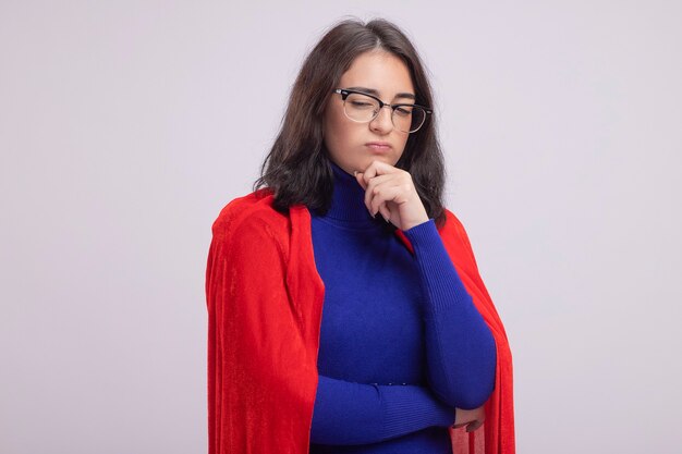 Jeune femme super-héros réfléchie en cape rouge portant des lunettes gardant la main sur le menton regardant vers le bas isolé sur un mur blanc avec espace de copie