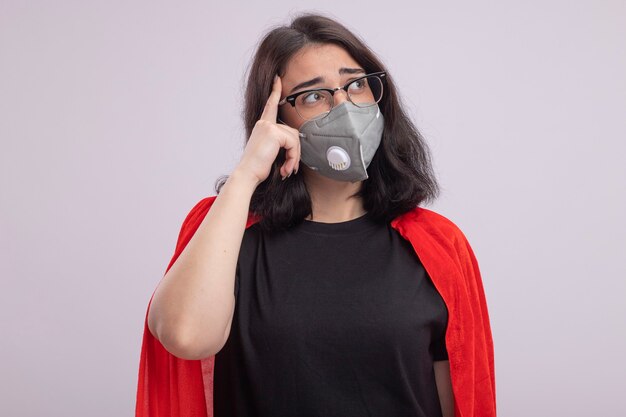 Jeune femme super-héros réfléchie en cape rouge portant des lunettes et un masque de protection regardant de côté faisant un geste de réflexion isolé sur un mur blanc