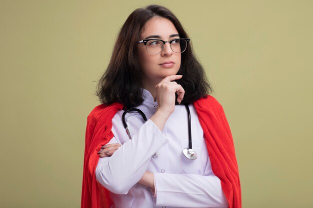 Jeune femme de super-héros réfléchie en cape rouge portant un uniforme de médecin et un stéthoscope avec des lunettes touchant le menton regardant le côté isolé sur un mur vert olive avec espace de copie