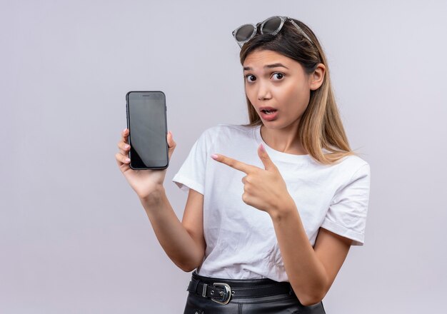 Une jeune femme surprenante en t-shirt blanc à lunettes de soleil montrant un téléphone mobile