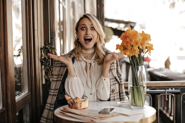 Une jeune femme surprise en blouse blanche et un manteau à carreaux en tweed est assise dans un café