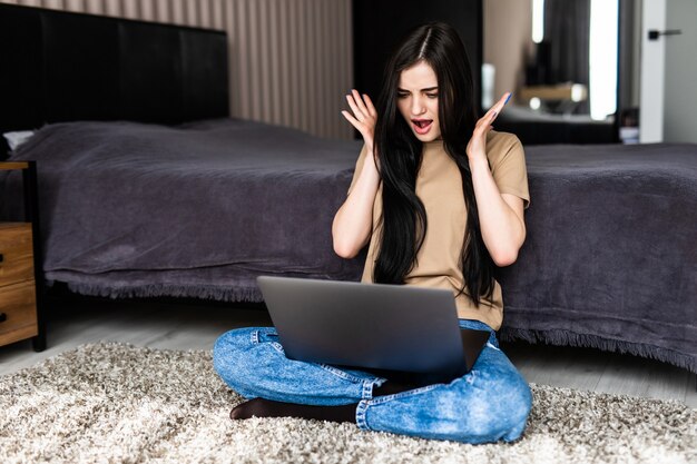 Jeune femme surprise choquée ayant un appel vidéo via un ordinateur portable allongé sur le sol dans un salon confortable pendant le travail à distance depuis la maison