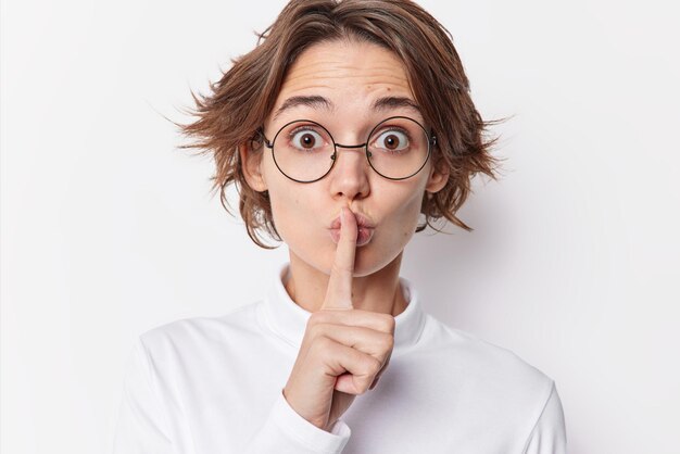 Une jeune femme surprise avec une coiffure à la mode semble abasourdie par la caméra qui appuie l'index sur les lèvres fait un geste de chut demande de se taire porte des lunettes rondes à l'intérieur. Ne laisse tomber aucun mot