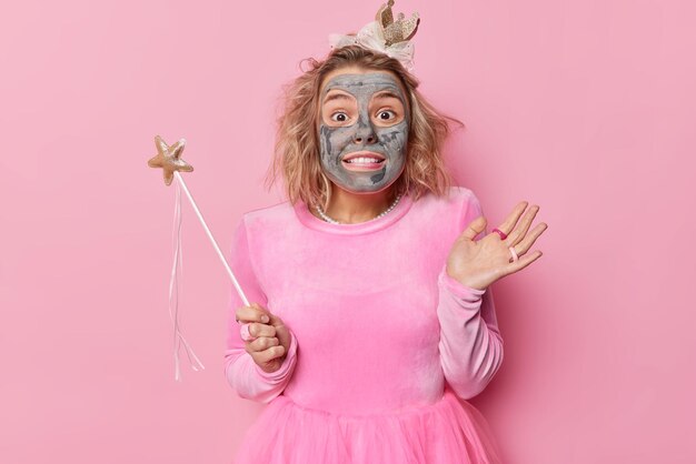 Une jeune femme surprise divertit les invités à la fête applique un masque d'argile de beauté porte une robe tient une baguette magique prête à réaliser vos souhaits et vos rêves isolés sur fond rose Fée magique à l'intérieur