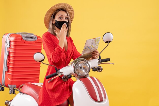 jeune femme surprise avec un masque noir tenant une carte près d'un cyclomoteur