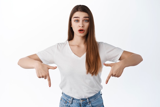 Jeune femme surprise pointant les doigts vers le bas, bouche ouverte étonnée, haussant les sourcils d'incrédulité, montrant quelque chose de nouveau, une bannière ou un logo, debout sur un mur blanc