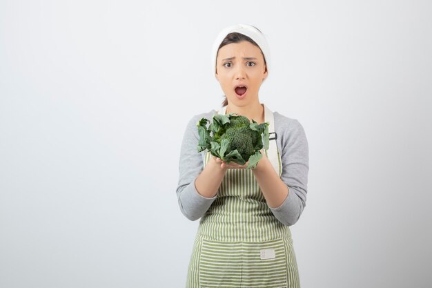 Jeune femme surprise en tablier tenant du brocoli frais sur blanc