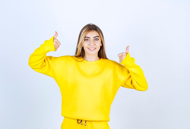 Jeune femme en survêtement jaune donnant les pouces vers le haut sur le mur blanc