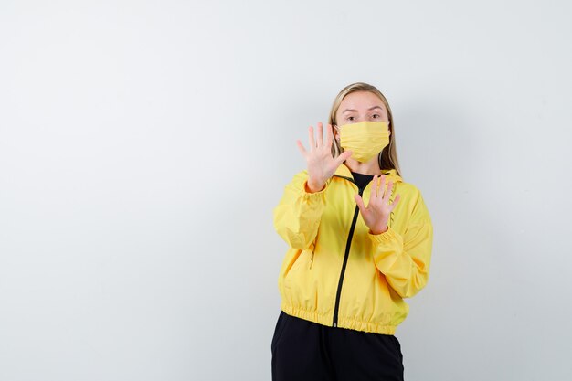 Jeune femme en survêtement, masque montrant le geste d'arrêt et l'air effrayé, vue de face.