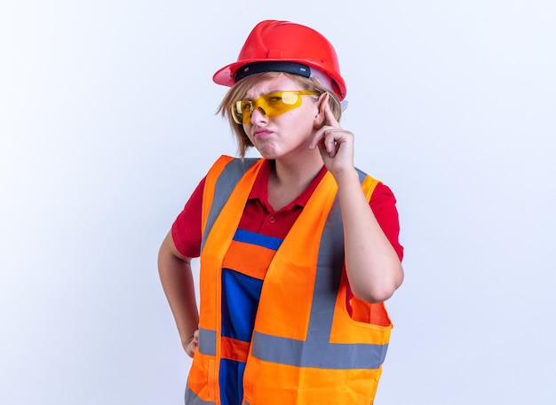 Jeune femme suspecte de constructeur en uniforme avec des lunettes montrant le geste d'écoute isolé sur fond blanc
