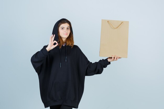 Jeune femme en sweat à capuche surdimensionné, pantalon montrant un geste correct tout en tenant le paquet et à la vue de face, confiant.