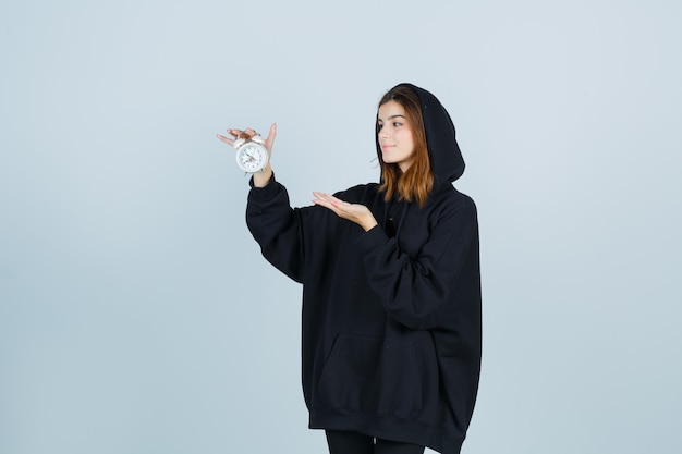 Jeune femme en sweat à capuche surdimensionné, pantalon tenant un réveil tout en faisant semblant de montrer quelque chose et à la vue de face positive.