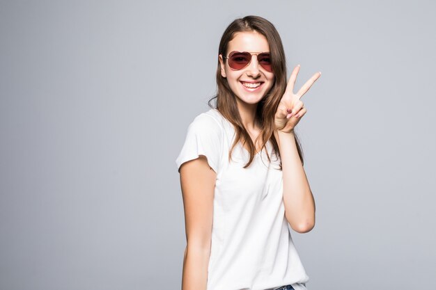 Jeune femme en t-shirt blanc et jean bleu montre le signe de la victoire devant un fond de studio blanc