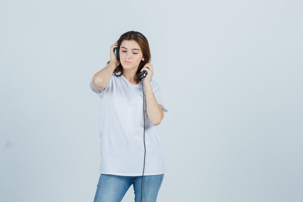 Jeune femme en t-shirt blanc, jeans décollant les écouteurs et regardant pensif, vue de face.