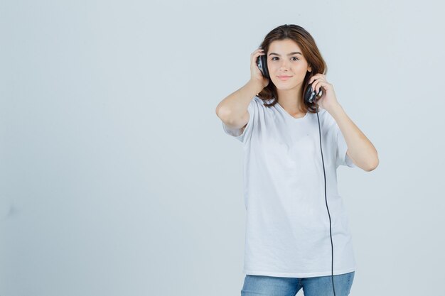 Jeune femme en t-shirt blanc, jeans décollant les écouteurs et à la rêveuse, vue de face.
