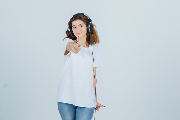 Jeune femme en t-shirt blanc, jeans montrant le pouce vers le haut tout en écoutant de la musique avec des écouteurs et à la vue de face, heureux.