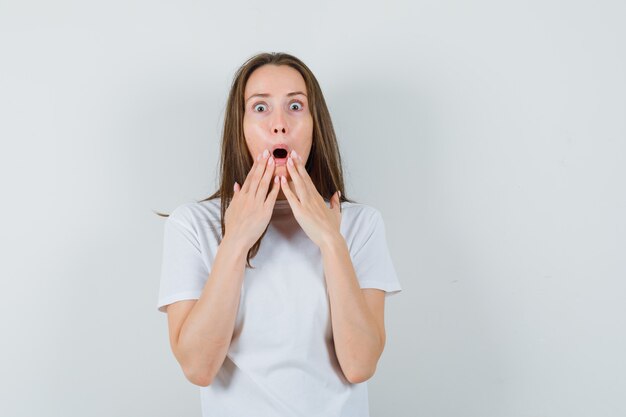 Jeune femme en t-shirt blanc, main dans la main près de la bouche ouverte et à la surprise