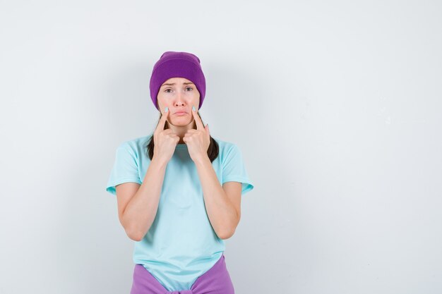 Jeune femme en t-shirt bleu, bonnet violet avec index près de la bouche, lèvres courbées et lugubre, vue de face.