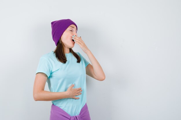 Jeune femme en t-shirt bleu, bonnet violet avec une main devant la bouche, une autre main sur le ventre en bâillant, et l'air endormi, vue de face.