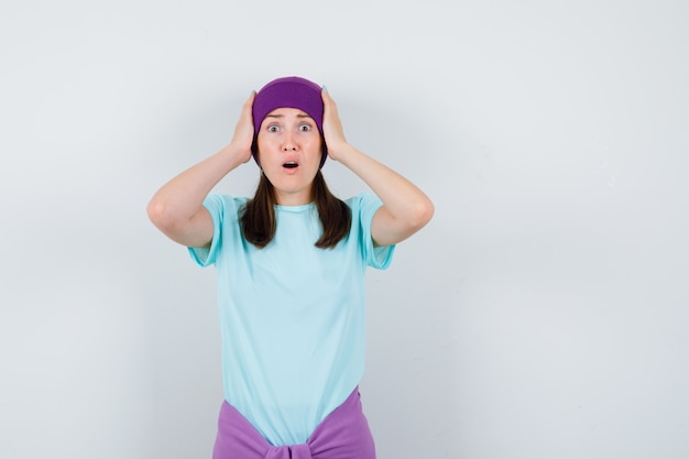 Jeune femme en t-shirt bleu, bonnet violet avec les mains sur la tête, gardant la bouche ouverte et l'air choqué, vue de face.