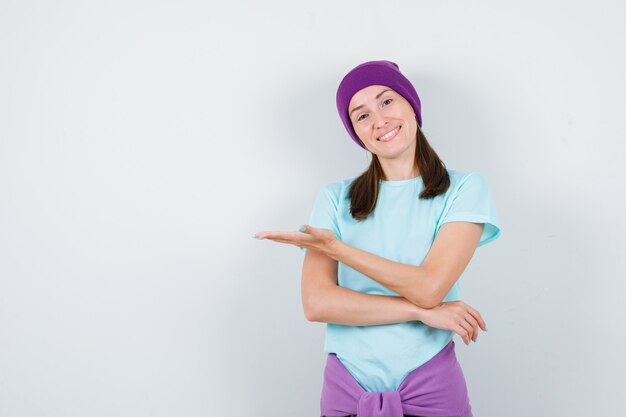 Jeune femme en t-shirt, bonnet montrant un geste de bienvenue et l'air joyeux, vue de face.