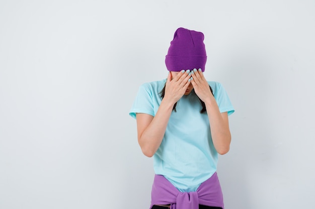 Jeune femme en t-shirt, bonnet penché la tête vers le bas, couvrant le visage avec les mains et l'air déprimé, vue de face.