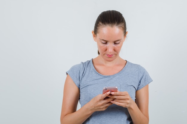 Jeune femme en t-shirt gris à l'aide de téléphone portable et à la vue de face, occupé.