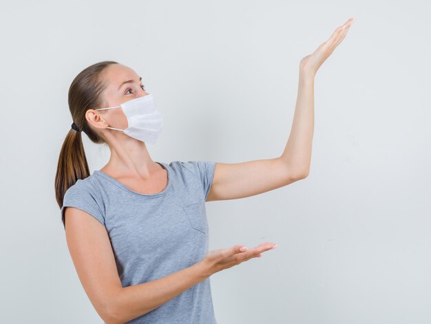 Jeune femme en t-shirt gris, masque montrant ou soulevant quelque chose et regardant gai, vue de face.