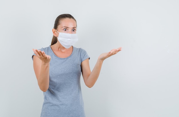 Jeune femme en t-shirt gris, masque soulevant les paumes dans le geste de questionnement, vue de face.