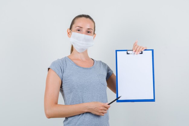 Jeune femme en t-shirt gris, masque tenant le presse-papiers et un stylo et à la joyeuse vue de face.