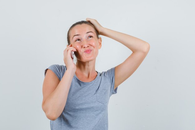 Jeune femme en t-shirt gris parlant au téléphone mobile et à la vue de face, hésitante.